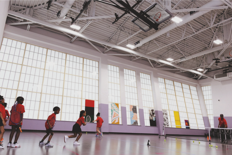 Children line up to play tennis at New Covenant Missionary Baptist Church's indoor tennis court, while Coach Tyrone Mason tosses the young athletes tennis balls. (Photo by Caeli Kean / South Side Weekly)